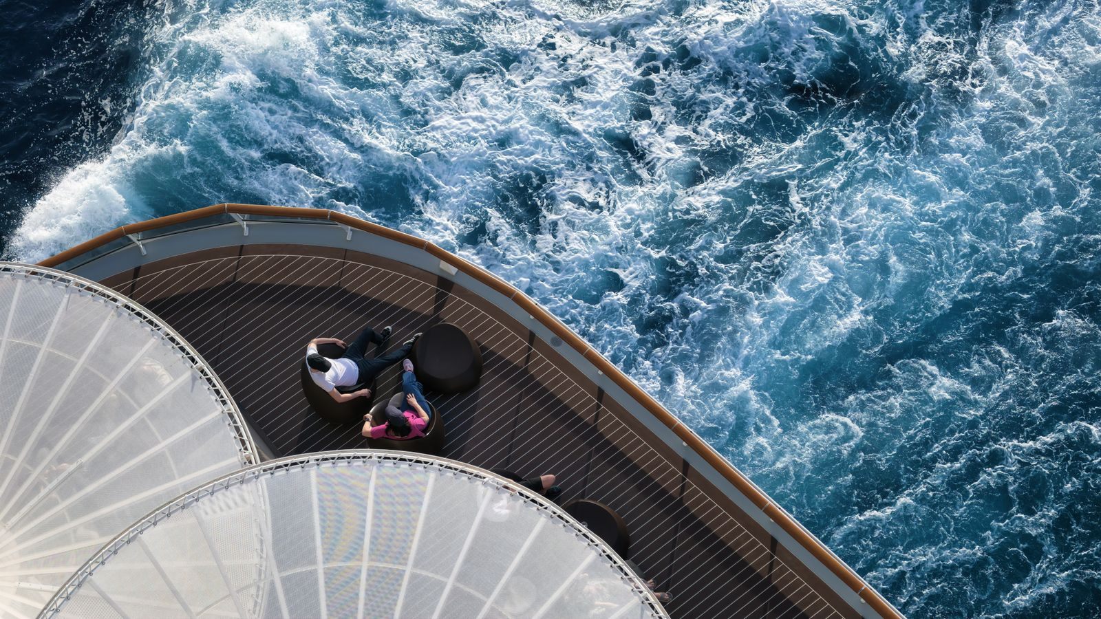 Two travelers relaxing on a curved stern deck above the churning wake of a cruise ship, the experience cruise marketing sells