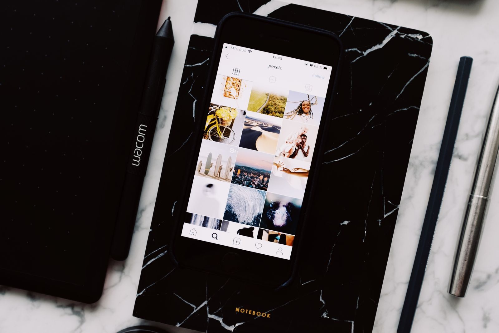 Smartphone showing a grid of travel photos on a marble desk next to a notebook, travel agent marketing ideas in progress